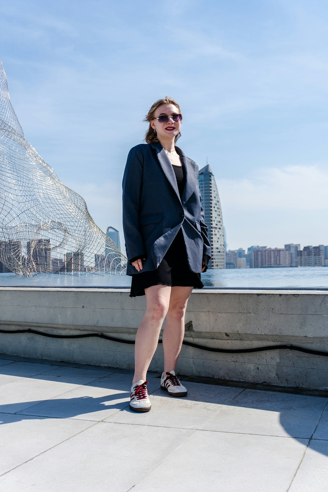 Woman in blazer standing by waterfront with cityscape background
