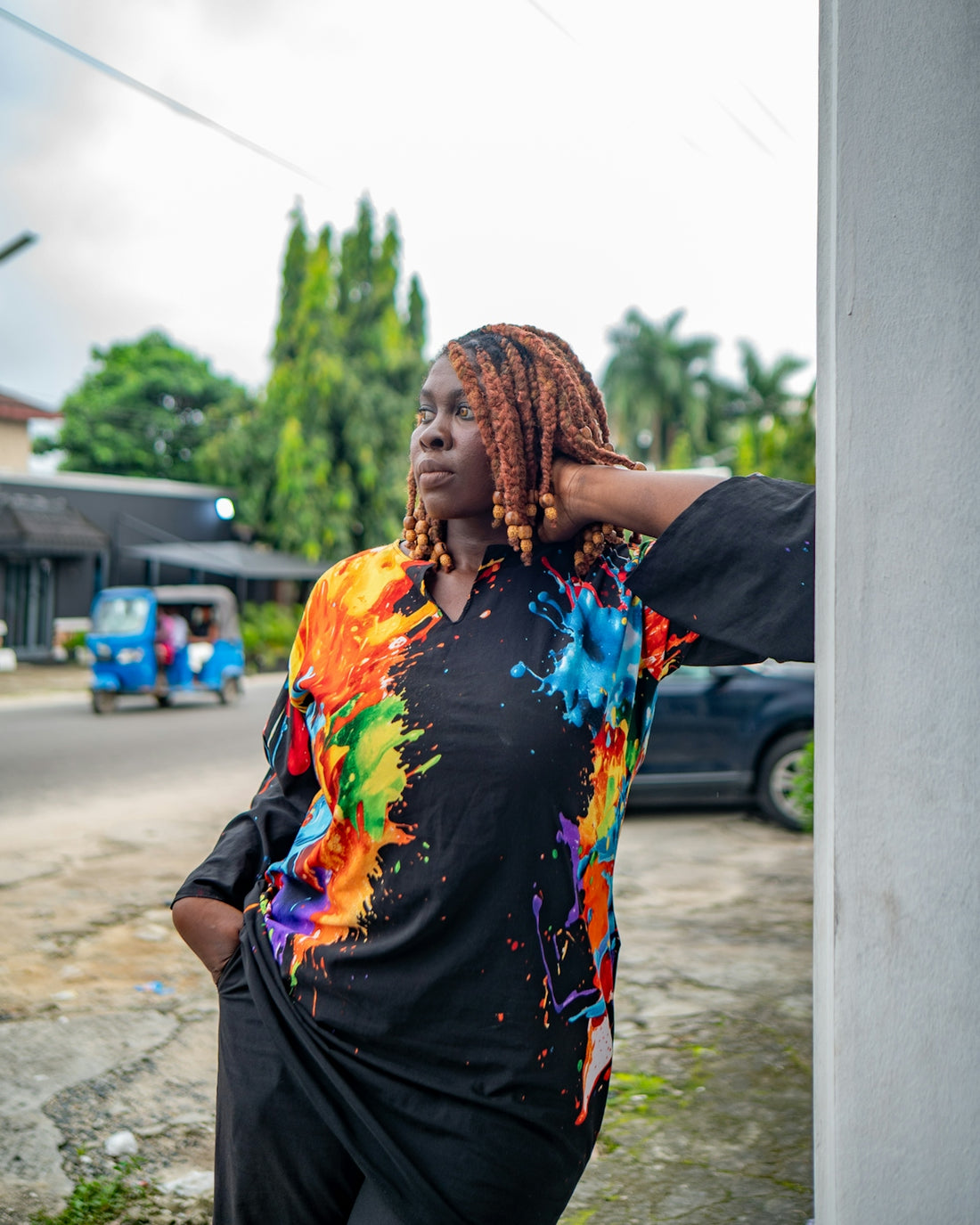 Woman in colorful paint-splattered shirt leans against wall.