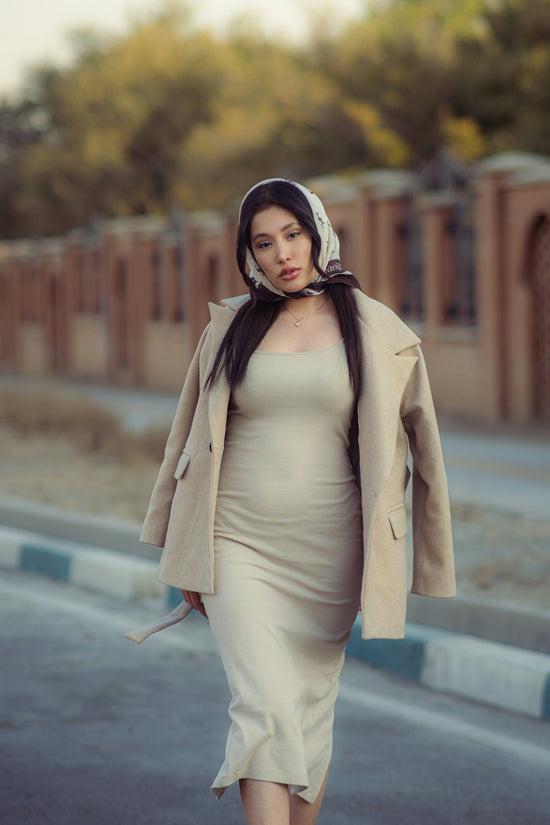 A woman in a beige dress and coat walks outdoors.