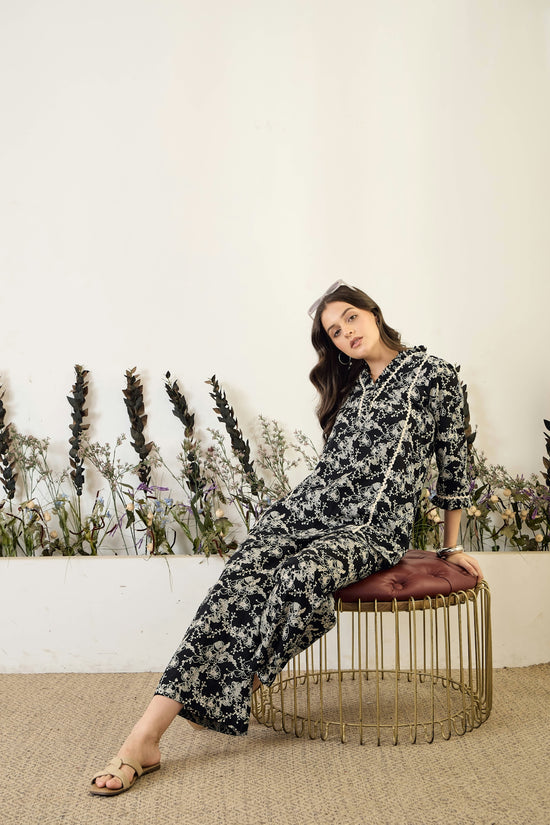Woman in floral jumpsuit sitting on stool.